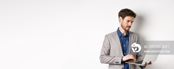 Handsome male entrepreneur working on laptop, looking serious at screen, standing against white background