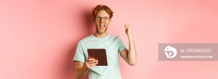 Image of happy redhead man triumphing, winning online with digital tablet and rejoicing, standing over pink background