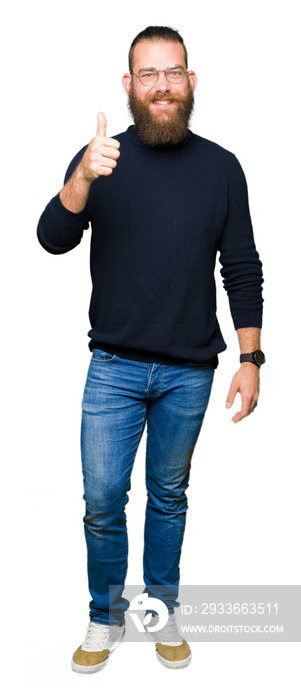 Young blond man wearing glasses and turtleneck sweater doing happy thumbs up gesture with hand. Approving expression looking at the camera showing success.