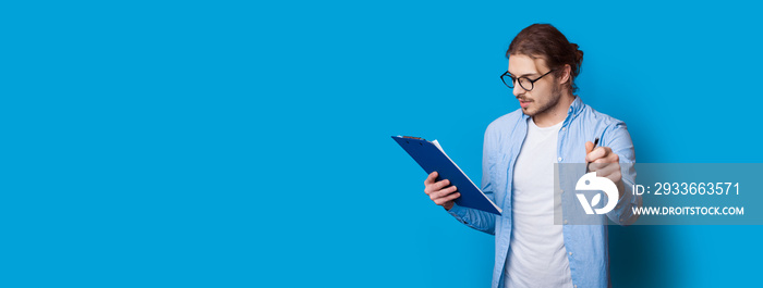 young businessman is posing on a blue wall with free space looking in a folder and holding a pen