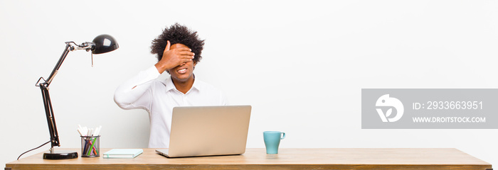 young black businessman covering eyes with one hand feeling scared or anxious, wondering or blindly waiting for a surprise on a desk