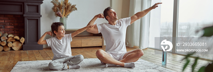 Merry little boy is training with shaply dad on mat at home and demonstrating muscles