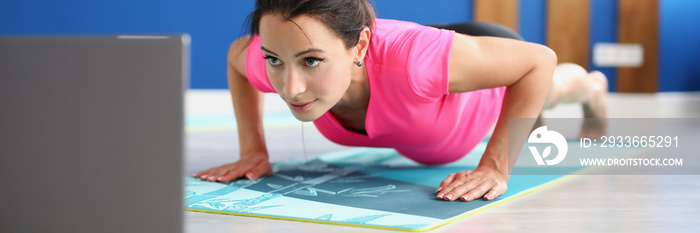 Woman in sportswear standing in plank on floor in front of laptop