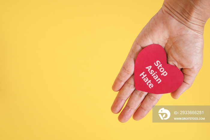 Stop Asian Hate , white message, on red heart paper in hand with yellow background and copy space , anti racist ,hate crimes against Asians concept