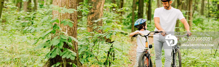 panoramic shot of father and son walking with bicycles in forest