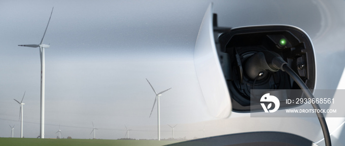 Close up of electric car inlet with a connected charging cable and double exposure with wind turbines