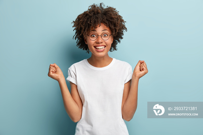 Overjoyed Afro American woman anticipates for victory, clenches fists, has broad smile, wears casual