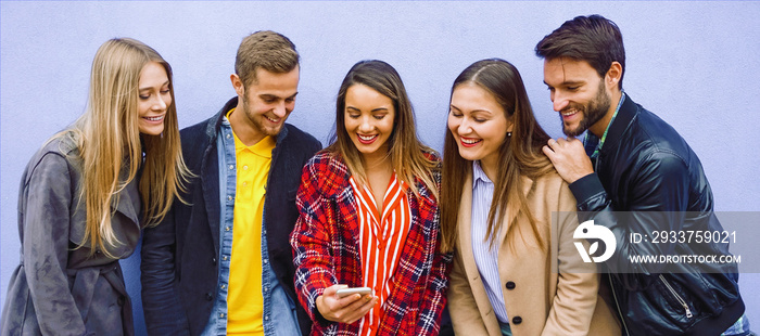 Young people standing by the wall while looking in smartphone and smiling- College friends checking 