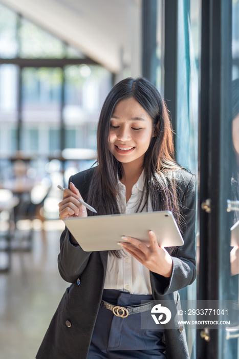 Beautiful Asian woman smiling standing near the window with a tablet at the office.