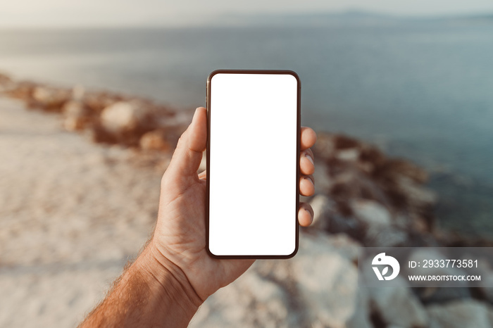 Man standing by the sea and holding mobile smart phone with blank mockup screen, closeup of hand