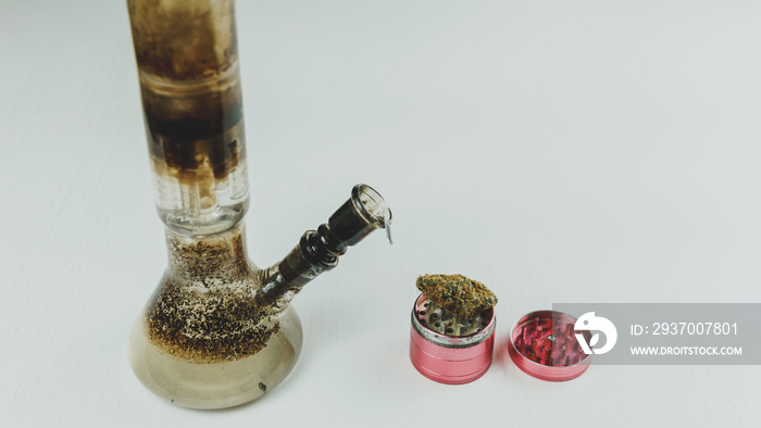 Close-up of dirty bong and grinder with medical marijuana buds on the white table. Smoking cannabis 