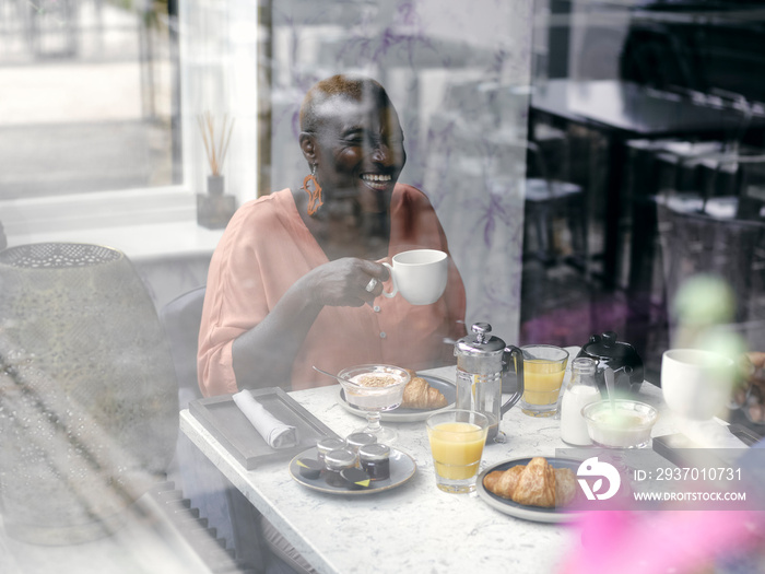 UK,ÊSmiling woman drinking coffee at breakfast table seen through window