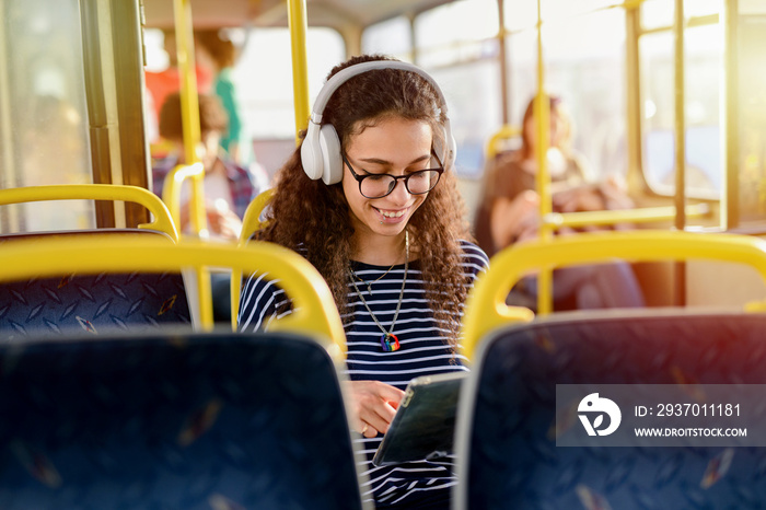 Beautiful young curly student girl sitting in a bus and listening to the music on her way to the sch