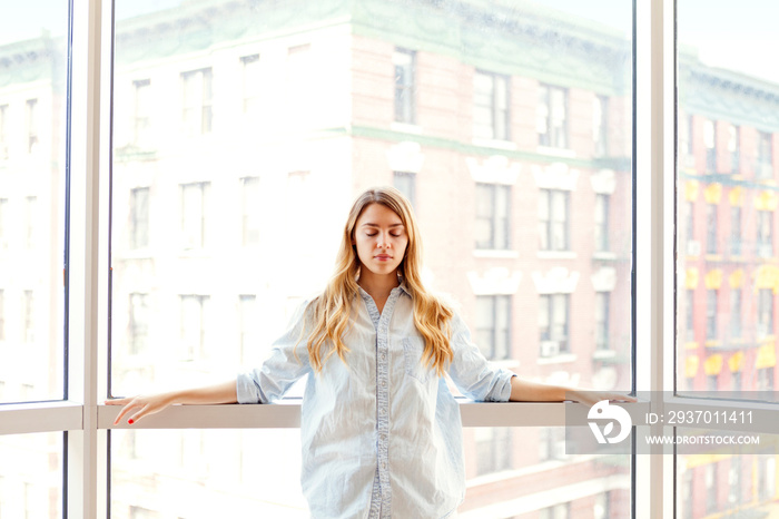 Young woman standing against window with closed eyes