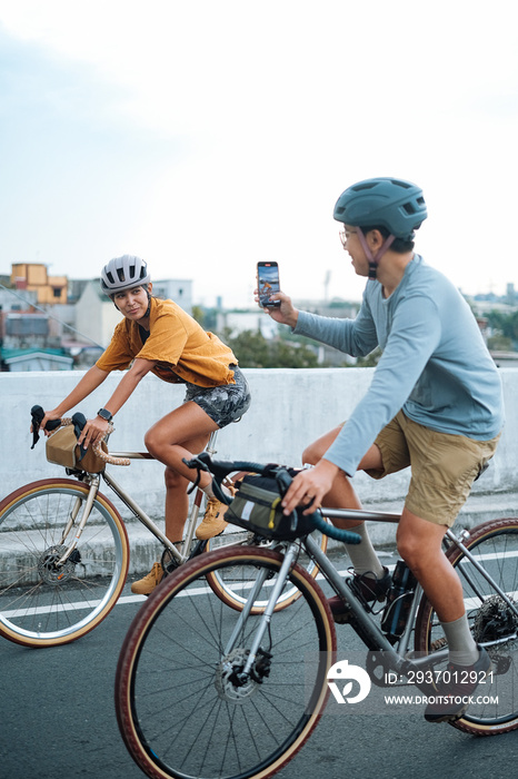 A young couple taking pictures with their mobile phones while on their bikes.