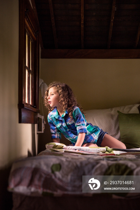 Girl (4-5) with curly hair on bed looking out through window