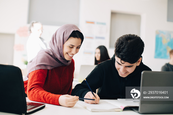 Happy male and female friends studying together at desk in classroom