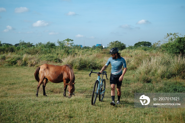 A young bearded cyclist is biking through a field with a horse