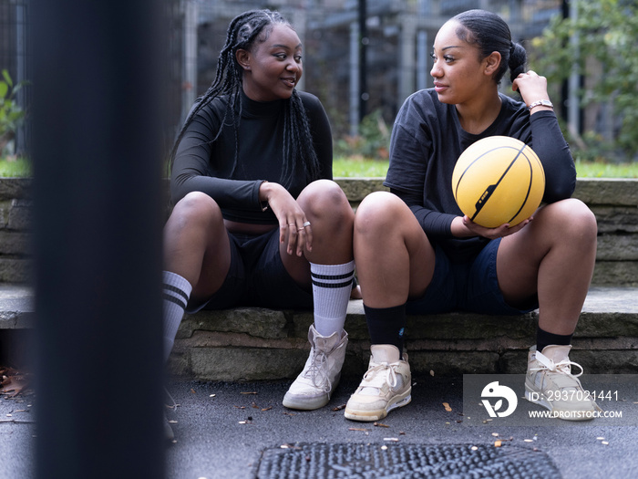 Two female friends sitting outdoors with basketball