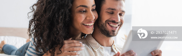 cheerful multiethnic couple looking at digital tablet, banner