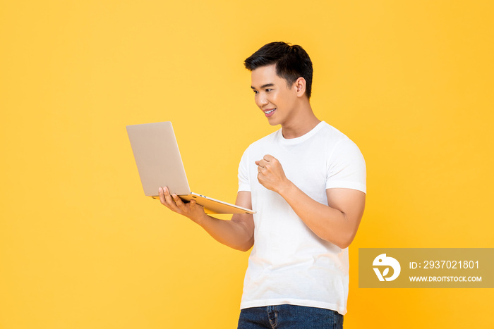 微笑的年轻英俊的亚洲男子在孤立的工作室里看着笔记本电脑，做出获胜的拳头手势