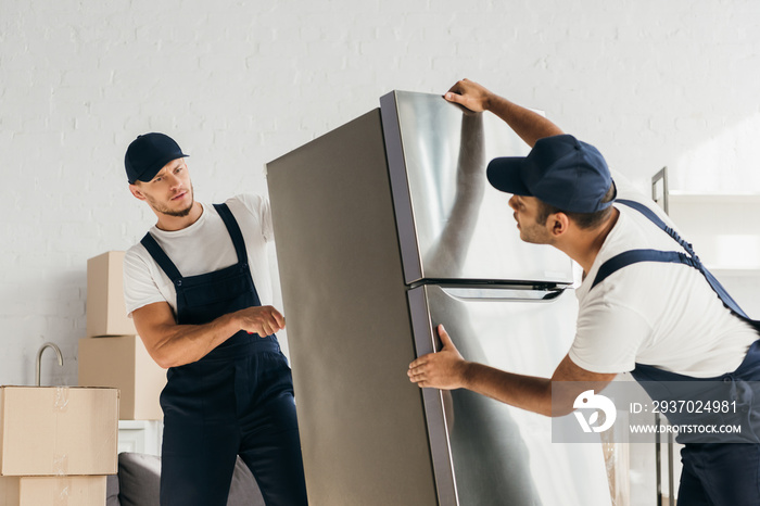 multicultural movers in uniform moving fridge in apartment