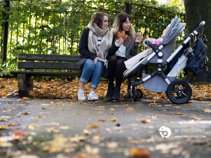 Lesbian couple sitting on bench next to a baby in stroller in park�