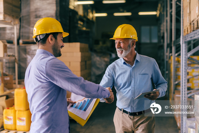 Two businessman with helmets on heads shaking hands for good finished job. Storage interior.