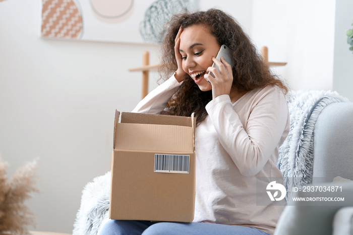 Happy young woman with parcel talking by mobile phone at home