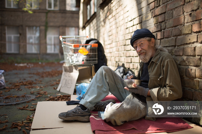 一个无家可归的老人坐在纸板上，手里拿着碗里的食物。如果流浪汉住在家里，午餐时间