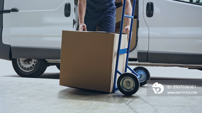 Cropped view of delivery man holding cardboard box near cart and auto outdoors