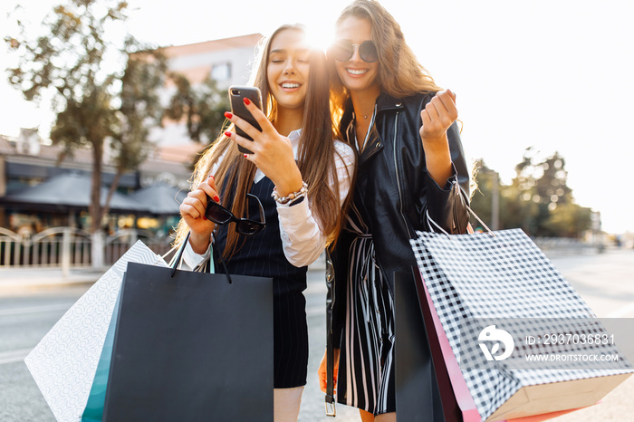 Beautiful young women in sunglasses hold shopping bags, use mobile phone and smile while standing on