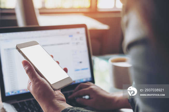 Mockup image of a woman holding mobile phone with blank white screen while using laptop with coffee 