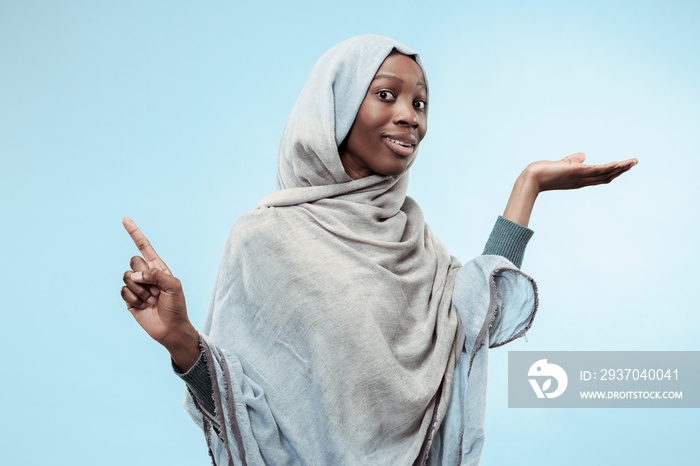 The beautiful young black african muslim girl wearing gray hijab at blue studio. She pointing to lef