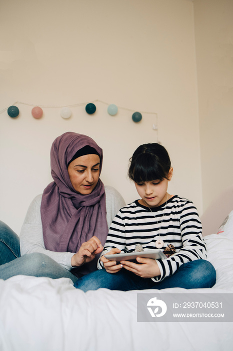 Mother explaining homework to daughter on digital tablet in bed at home