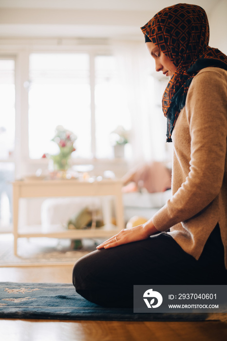 Full length of woman praying with closed eyes while sitting on carpet at home