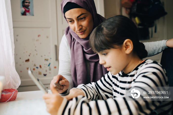 Mature mother assisting girl in homework while using digital tablet at home