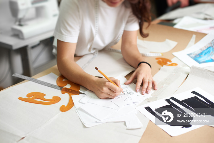 Young smiling female designer drawing pencil sketches of clothing sitting at big table with flat pap