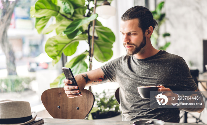 Handsome hipster man relaxing using digital smartphone with coffee and looking at screen typing mess