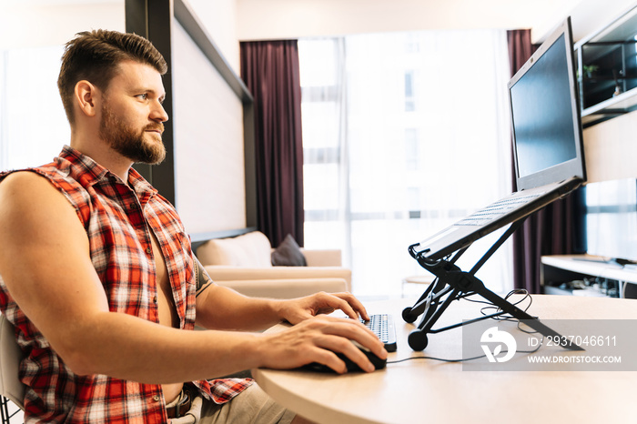 Man with an open checkered sleeveless shirt working remotely in front of a laptop using the mouse