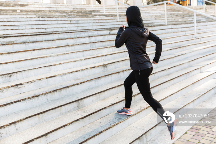 Young muslim sports fitness woman dressed in hijab and dark clothes running outdoors at the street.