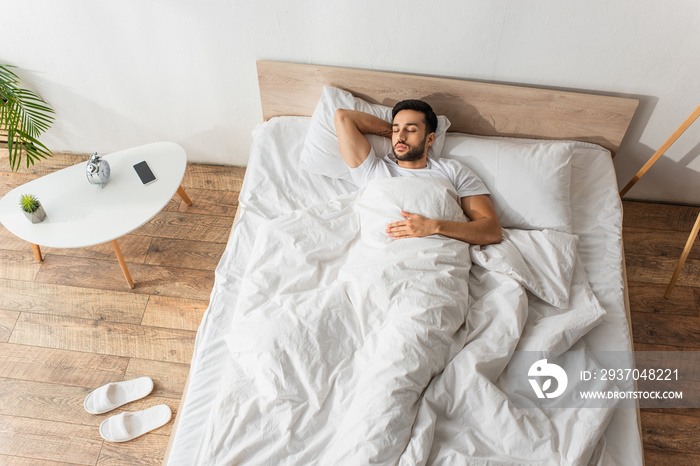 Top view of bearded man sleeping on white bedding near smartphone and alarm clock