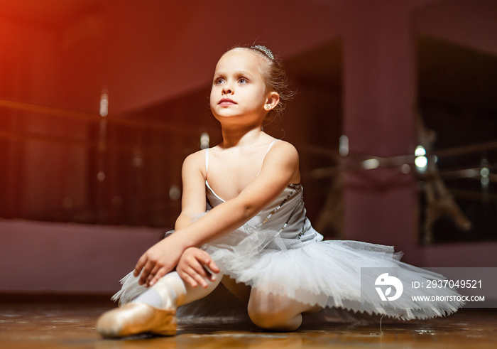 Little ballerina in white tutu sitting on floor and resting in dance studio. Pretty small girl in ba