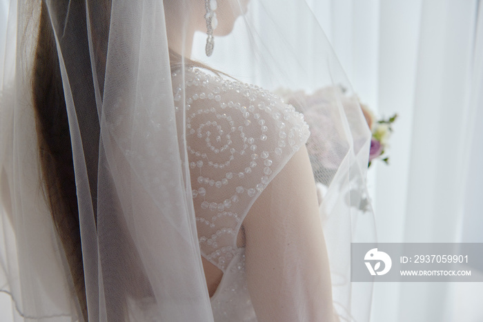穿着婚纱的美丽新娘拿着新娘花束站在房间窗户附近的肖像，复制品