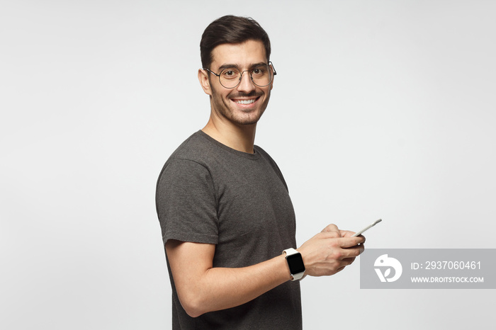 Young man standing isolated on gray background, holding smartphone, looking at camera and smiling ni