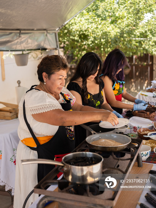 Right view image of an Indigenous mother putting seeds in a pan alongside her daughters who are dese