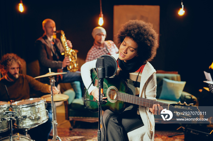 Mixed race woman singing and playing guitar while sitting on chair with legs crossed. In background 