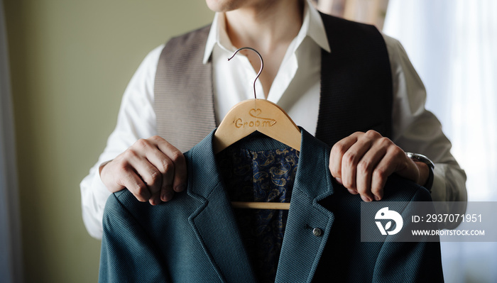 Groom in a white shirt and vest holds a jacket on a hanger