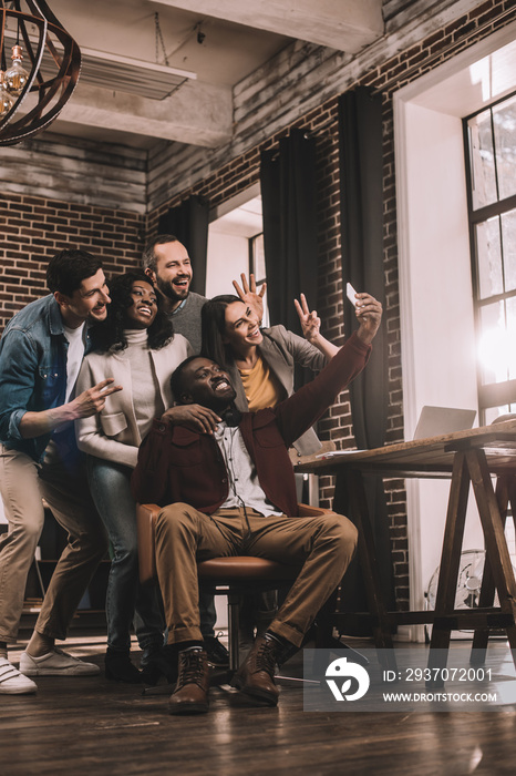 group of cheerful multiethnic coworkers taking selfie using smartphone in loft office with backlit