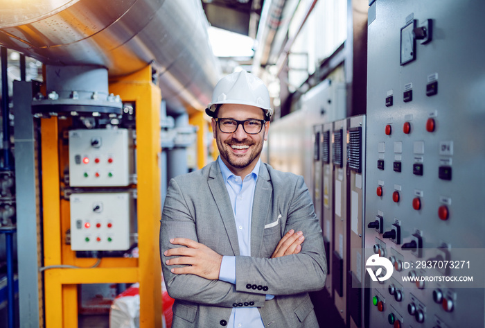 Smiling handsome caucasian businessman in gray suit and with helmet on head standing with arms folde